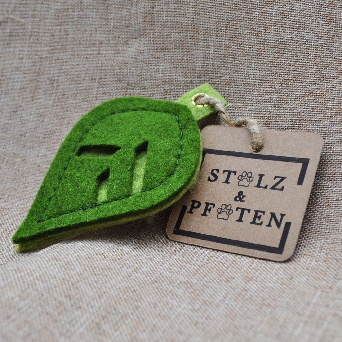Schlüsselanhänger in Blattform aus Merino-Wollfilz mit Produktanhänger aus Karton, befestigt an einer Hanfschnur, auf beigem Leinenstoff