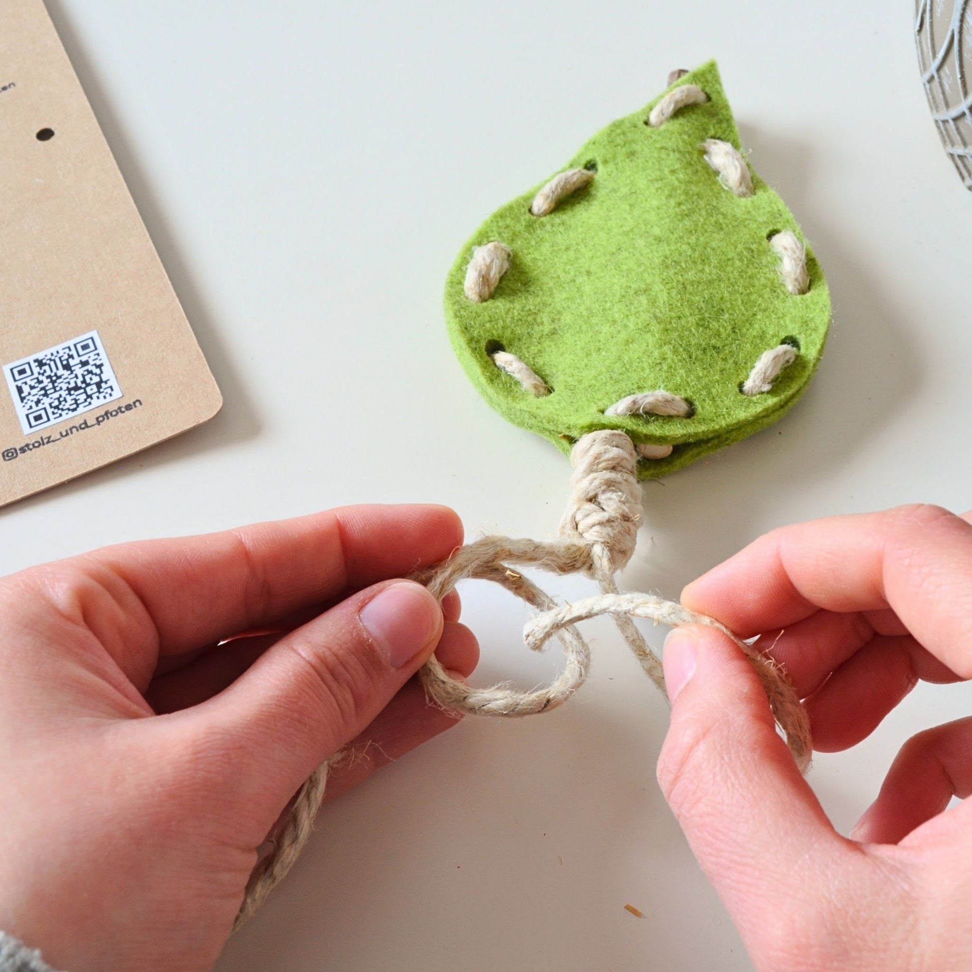 Pfotenwerk DIY-Katzenspielzeug beim Knoten der Hanfschnur, Hände arbeiten am Filzblatt, Anleitung liegt daneben